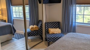 Ein Hotelzimmer mit einem großen Bett, einer Sitzecke mit einem gemusterten Sessel, einem Spiegel und einem an der Wand befestigten Fernseher.