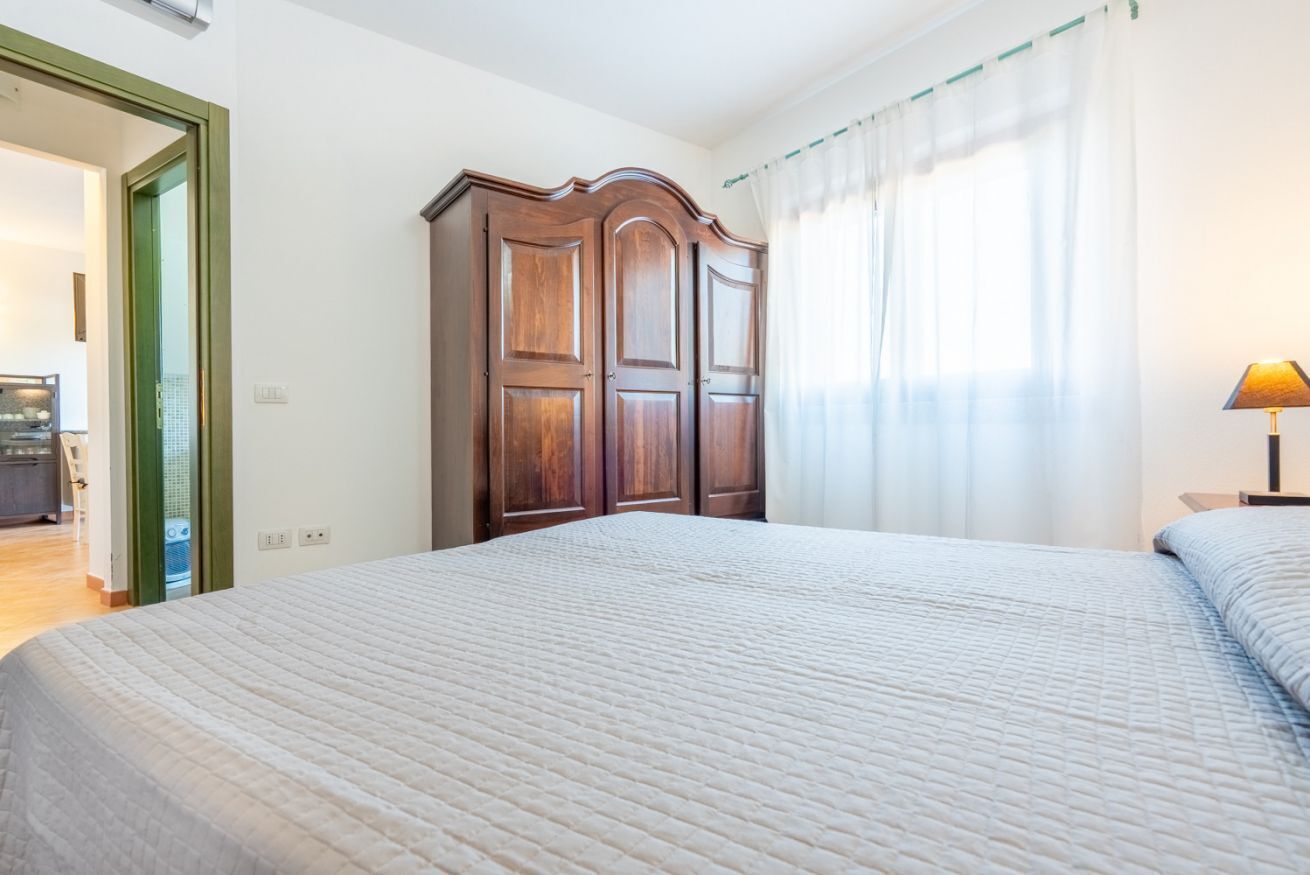 A bedroom with a large bed, a wooden wardrobe, and a window with sheer curtains.