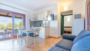 Une cuisine moderne avec des meubles blancs, un coin repas comprenant une table et des chaises, un téléviseur à écran plat fixé au-dessus du comptoir, et une porte-fenêtre coulissante donnant sur un espace extérieur.
