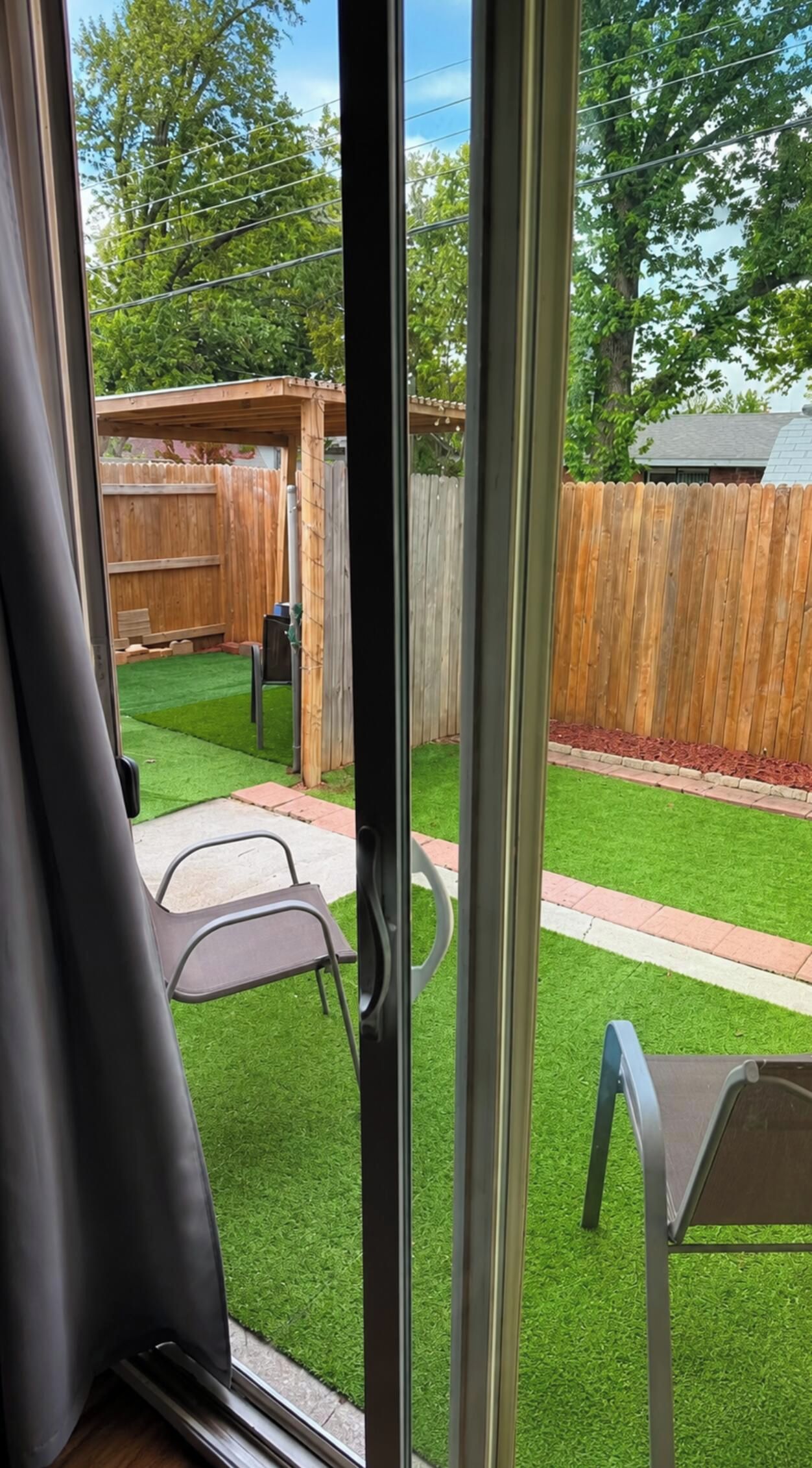 Vista per una porta corredissa d'un jardí amb gespa artificial, una tanca de fusta i una zona de pati amb cadires.