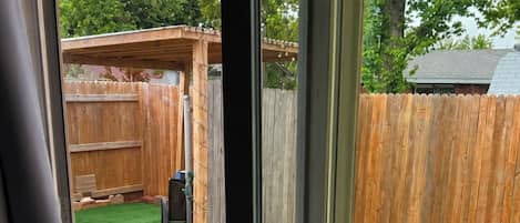 En vy genom en skjutdörr i glas som vetter ut mot en trädgård med konstgräs, ett trästaket och en uteplats med stolar.