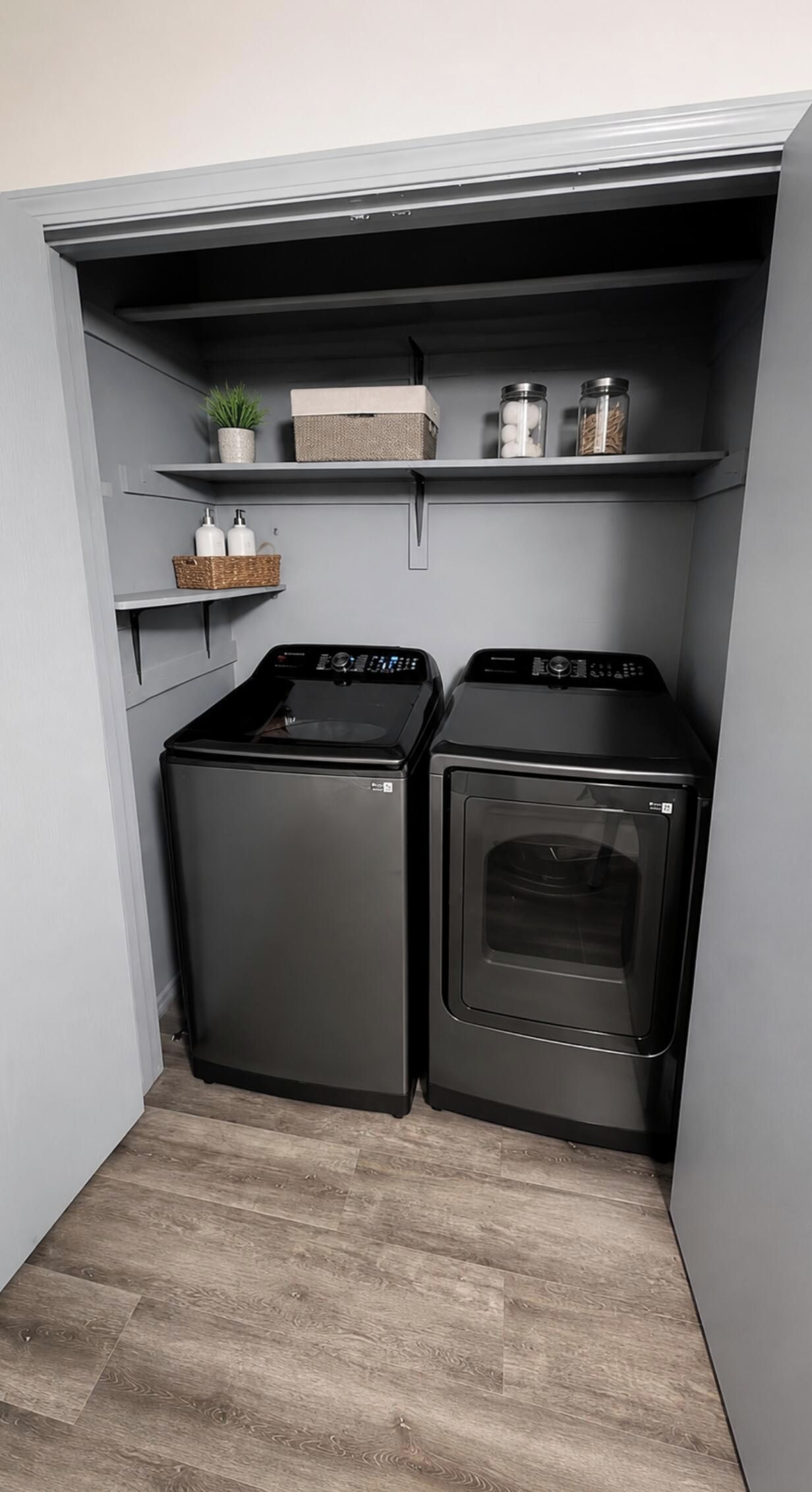 Une buanderie équipée de deux machines à laver à chargement frontal et d’un sèche-linge, d’une petite étagère avec une plante et des boîtes de rangement, et d’un mur gris.