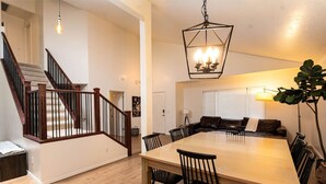 Amplia sala de estar con una mesa de comedor de madera, sillas negras, una planta grande en maceta y una escalera con barandillas de madera.