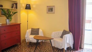 A room with a patterned floor, a red dresser, two white chairs with cushions, a small table, a floor lamp, and a framed picture on the wall. - To complete (Catania)