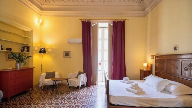 A hotel room with a bed, a chair, a desk, and a shelf. - To complete (Catania)