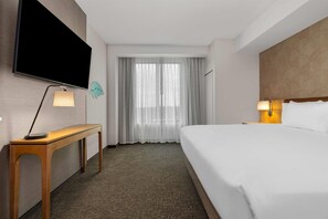 A hotel room with a bed, a desk with a lamp, a TV mounted on the wall, and a window with sheer curtains. - Drury Plaza Hotel Tampa Brandon (Tampa)