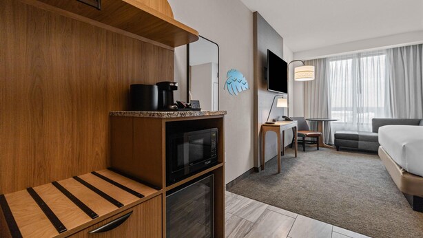 A hotel room with a wooden bench, a microwave, a small table with a lamp, a TV, a sofa, and a large window with curtains. - Drury Plaza Hotel Tampa Brandon (Tampa)