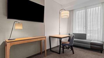 A hotel room with a flat-screen TV, a desk with a lamp, a chair, a sofa, and a large window with sheer curtains.