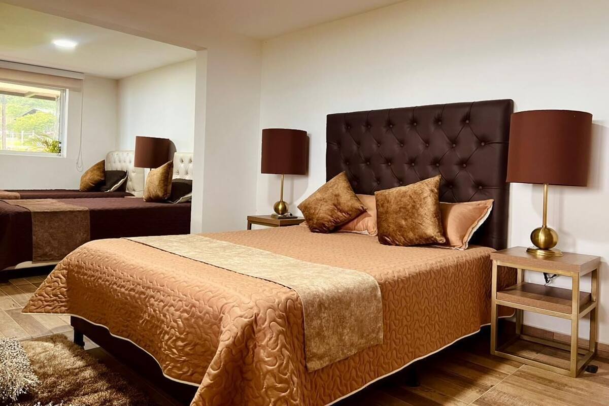 Un dormitorio con una cama grande, dos mesitas de noche con lámparas, una mesita auxiliar y una ventana con vistas.