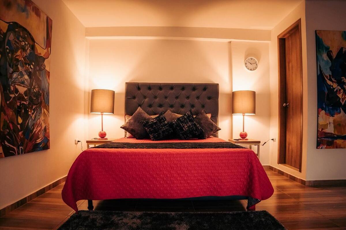 Un dormitorio con cabecera capitonada, colcha roja, dos lámparas de noche y un reloj en la pared.