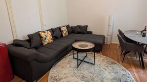 Una sala de estar moderna con un sofá seccional gris oscuro, una mesa de centro redonda, un tapete estampado y un comedor con una mesa pequeña y sillas.