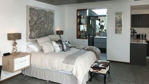 A bedroom with a bed, bedside tables, a nightstand, a bench, a large abstract painting, a mirror, and a kitchenette. - Charming garden suite (Belmont)