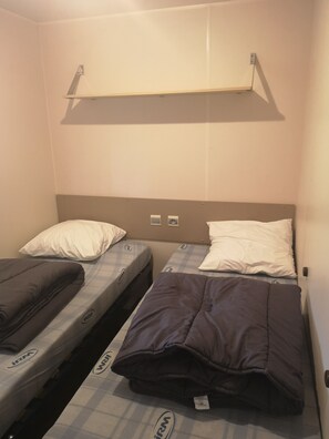 Quarto de hotel pequeno e individual, com duas camas de solteiro, cada uma com travesseiro e cobertor. Há uma prateleira sobre as camas e uma tomada na parede.