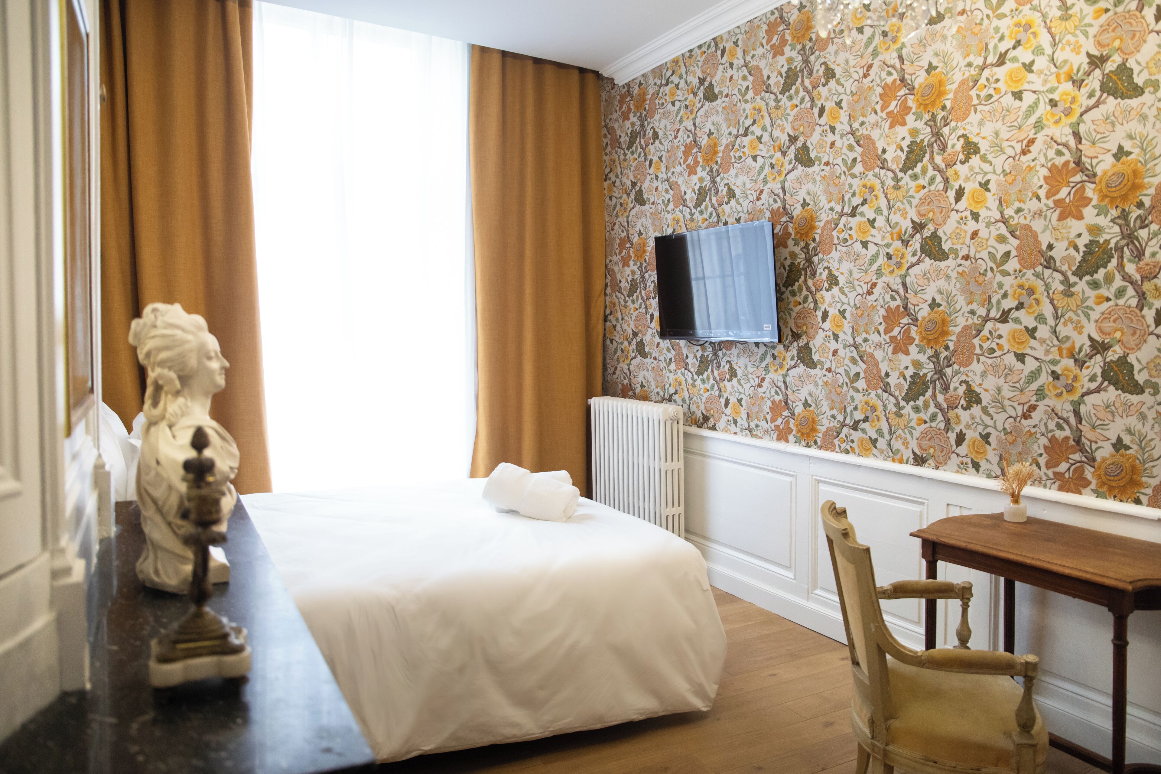 A hotel room with a bed, a wooden chair, a TV mounted on the wall, and a floral wallpaper.