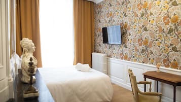 A hotel room with a bed, a wooden chair, a TV mounted on the wall, and a floral wallpaper.