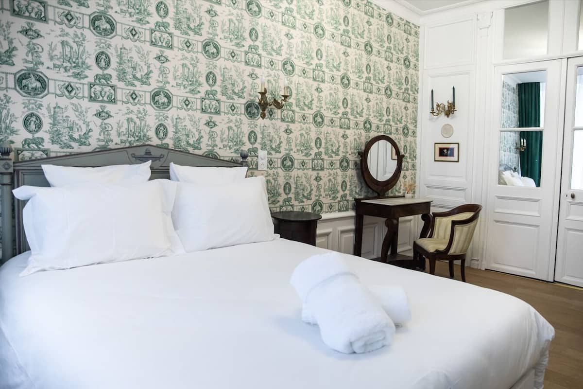 A neatly made bed with white linens, a round mirror, a small wooden table, and a chair in a room with patterned wallpaper and white walls.