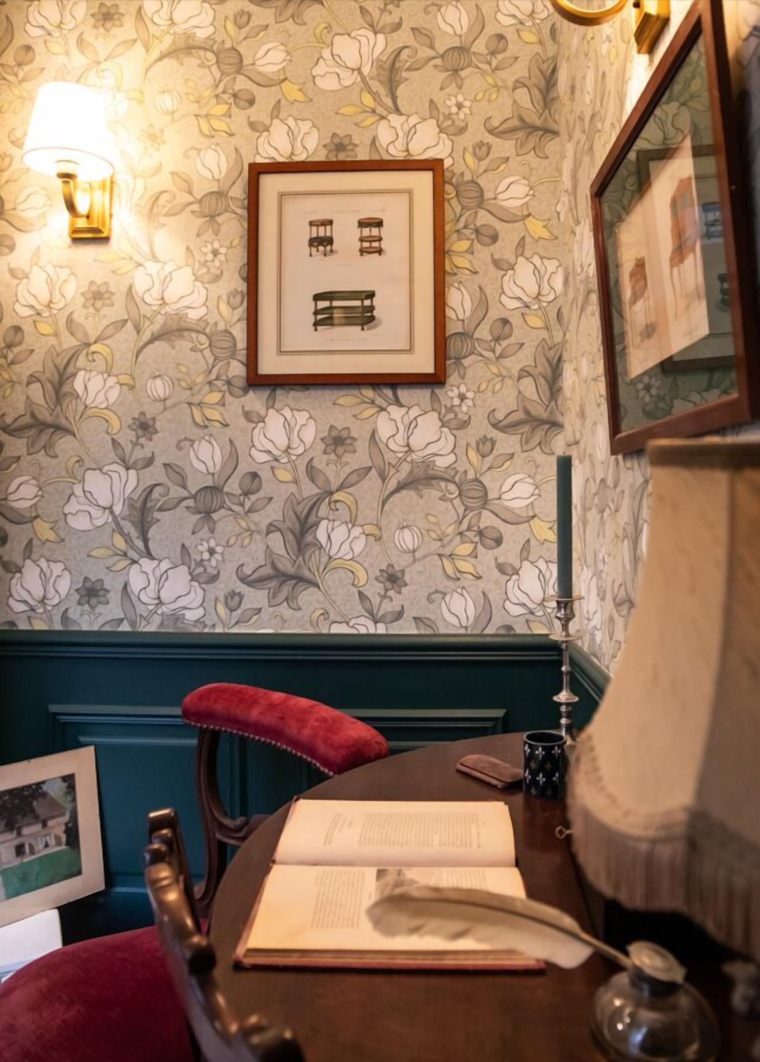 Une pièce avec un papier peint à motifs floraux, un tableau encadré représentant des meubles, un fauteuil rouge rembourré, une table avec un livre ouvert, une lampe et un miroir encadré.