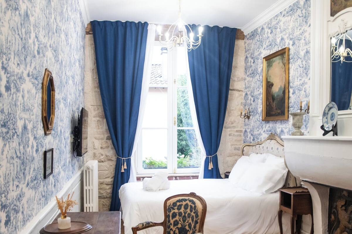 A room with a bed, a chair, a table, a chandelier, and a window with blue curtains.