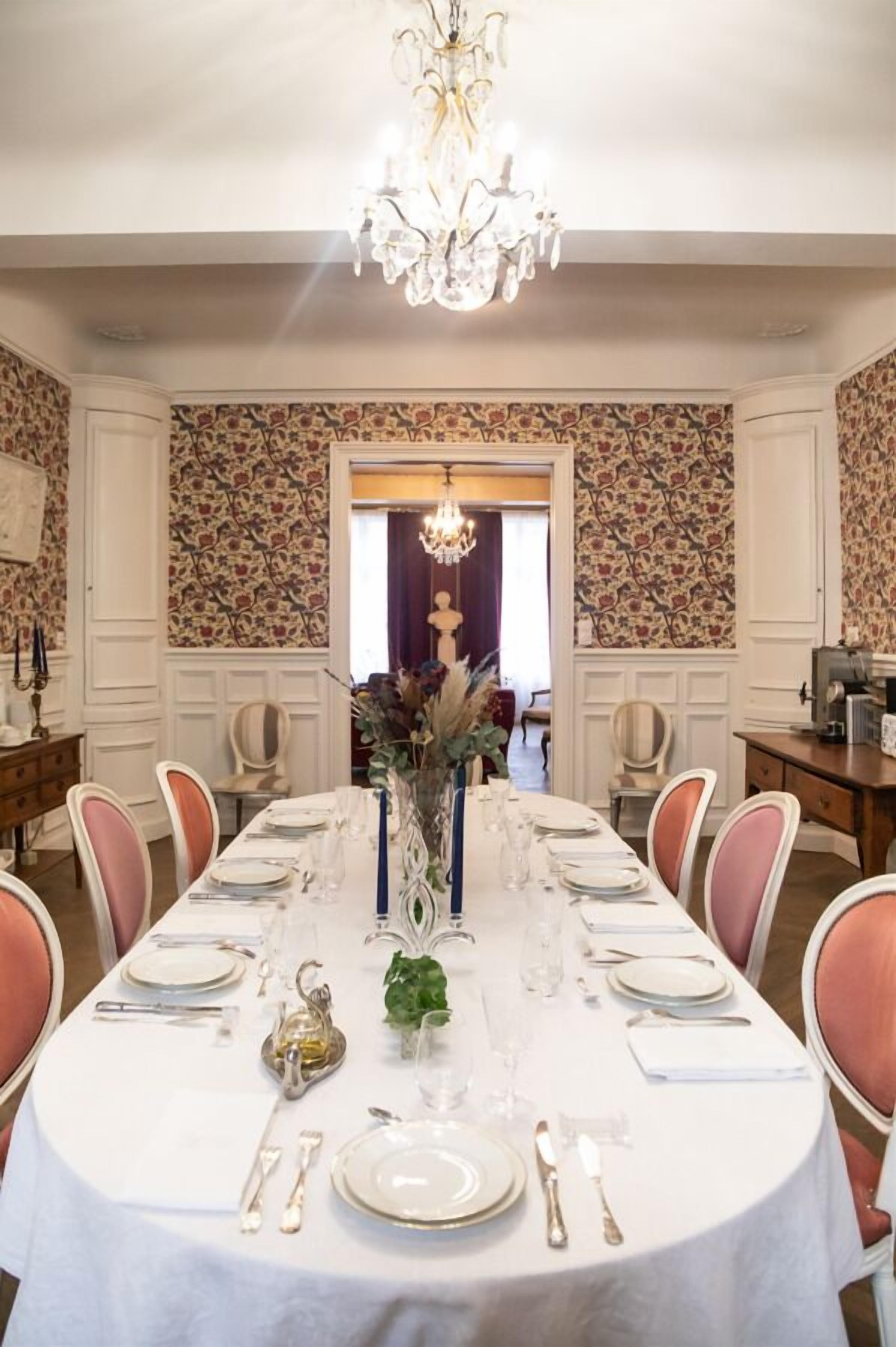 A dining room with a white table set for six, surrounded by pink chairs. A chandelier hangs from the ceiling, and the walls are decorated with floral wallpaper.