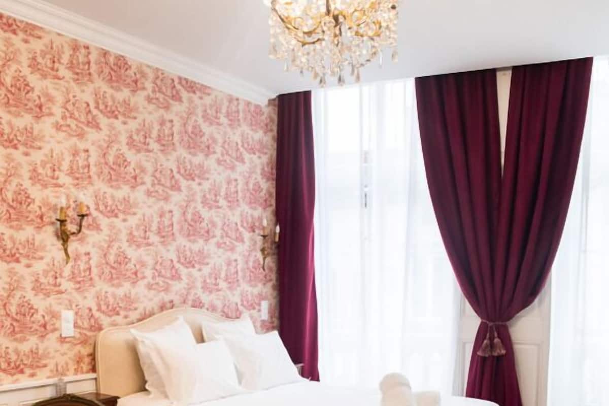A bedroom with a large bed, wooden floor, a chandelier, and patterned wallpaper.