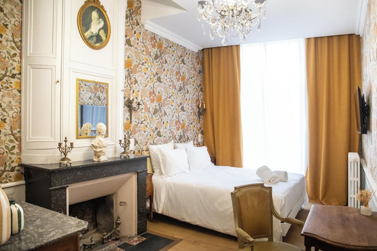 A hotel room with a bed, a desk with a mirror, a chandelier, and floral wallpaper.