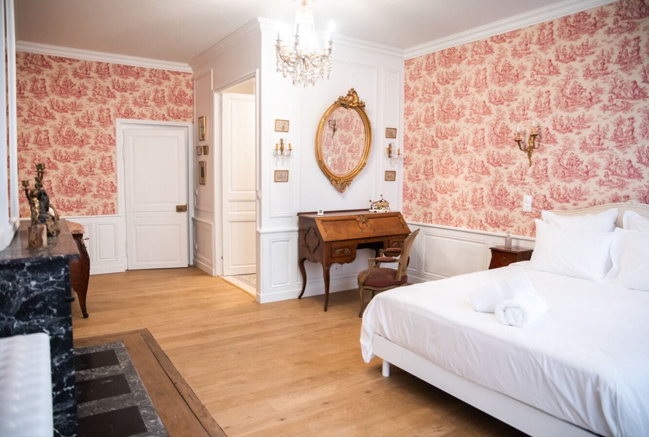 Quarto de hotel com piso de madeira, cama branca, escrivaninha de madeira, espelho e lustre.