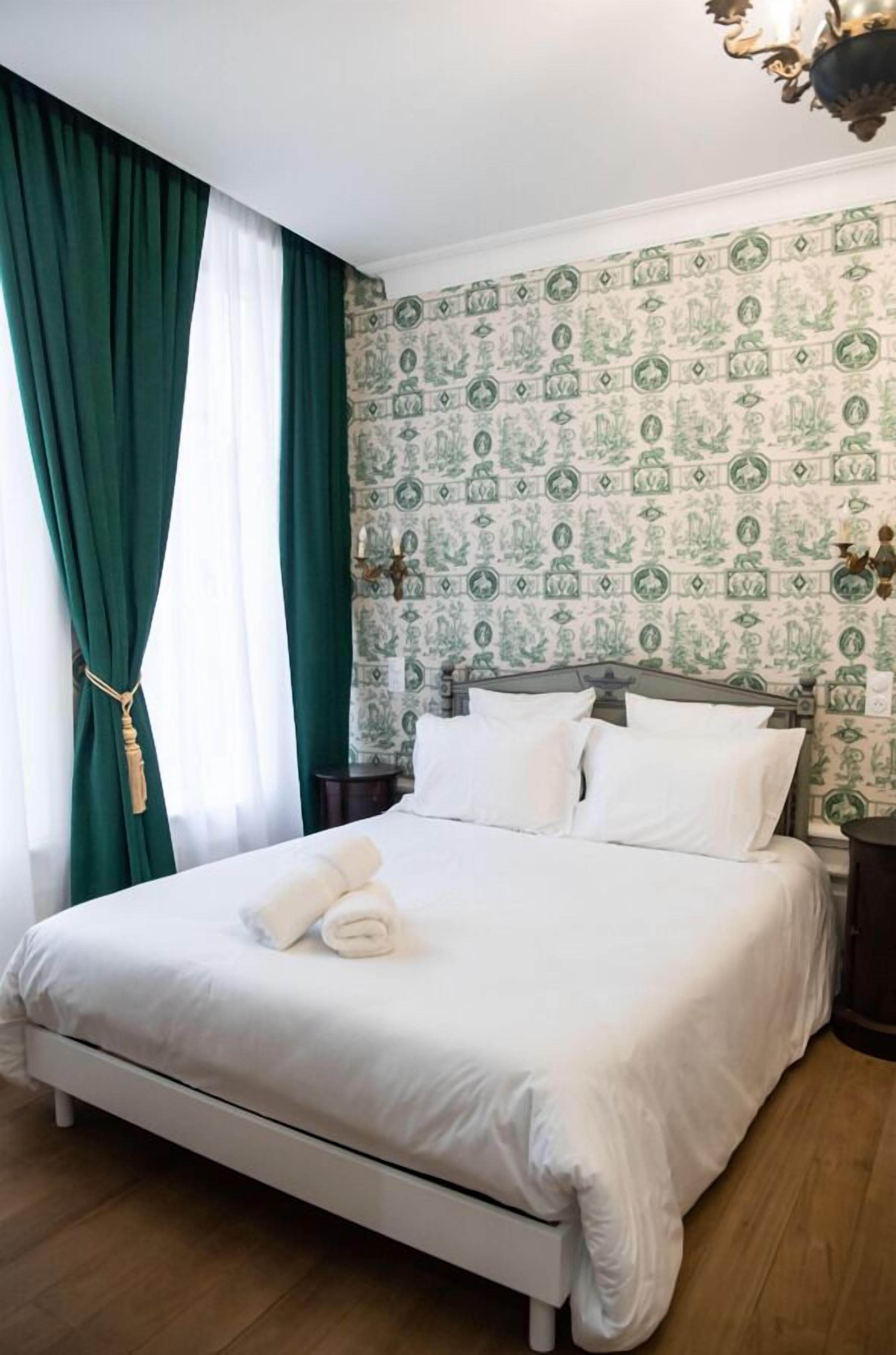Una cama bien hecha con sábanas blancas y una toalla enrollada, una mesita de noche con una lámpara y un papel tapiz estampado.