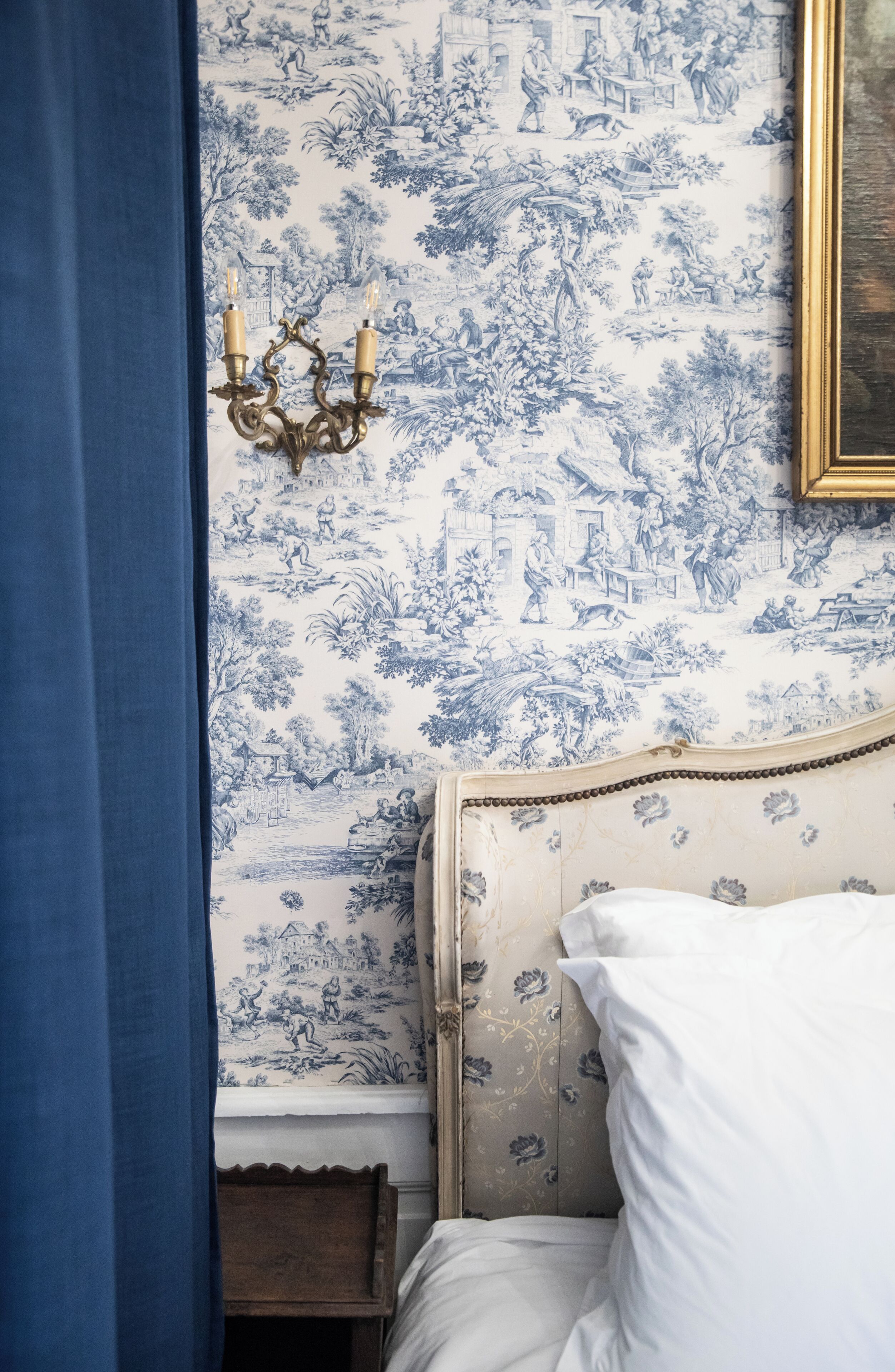 Un dormitorio con una cama, una cortina azul, un papel tapiz floral azul y blanco en la pared, y un candelabro dorado.