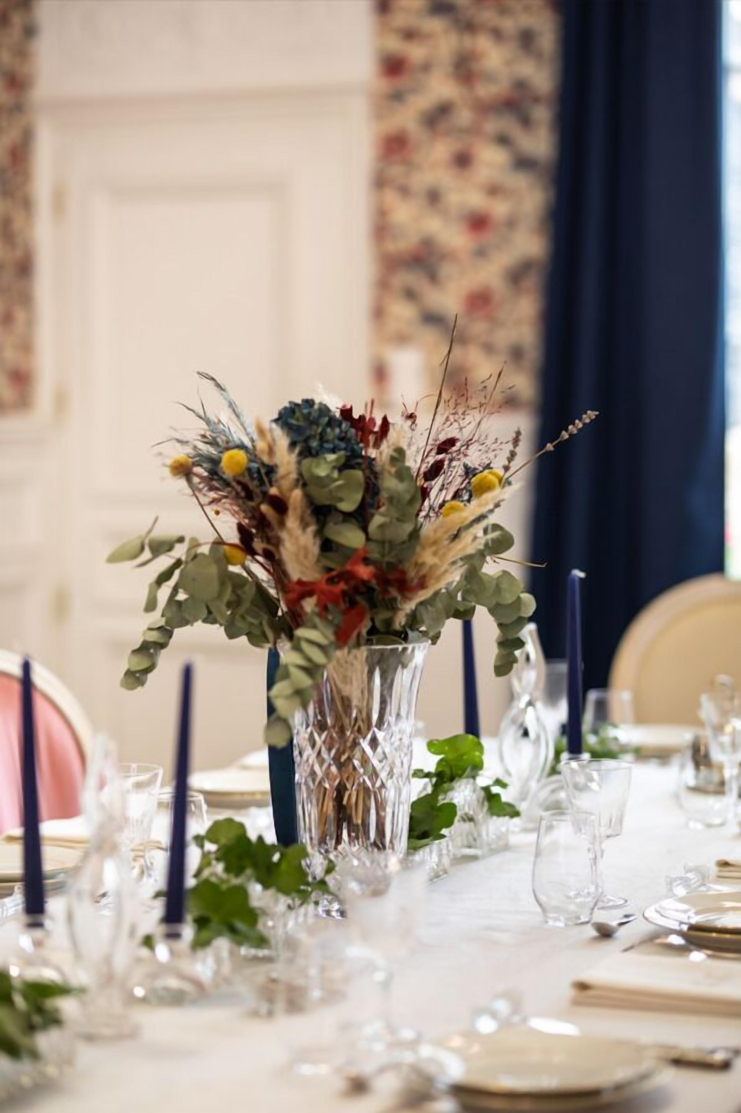 A dining table set with clear glassware, a decorative vase with dried flowers, and a dark blue curtain in the background.