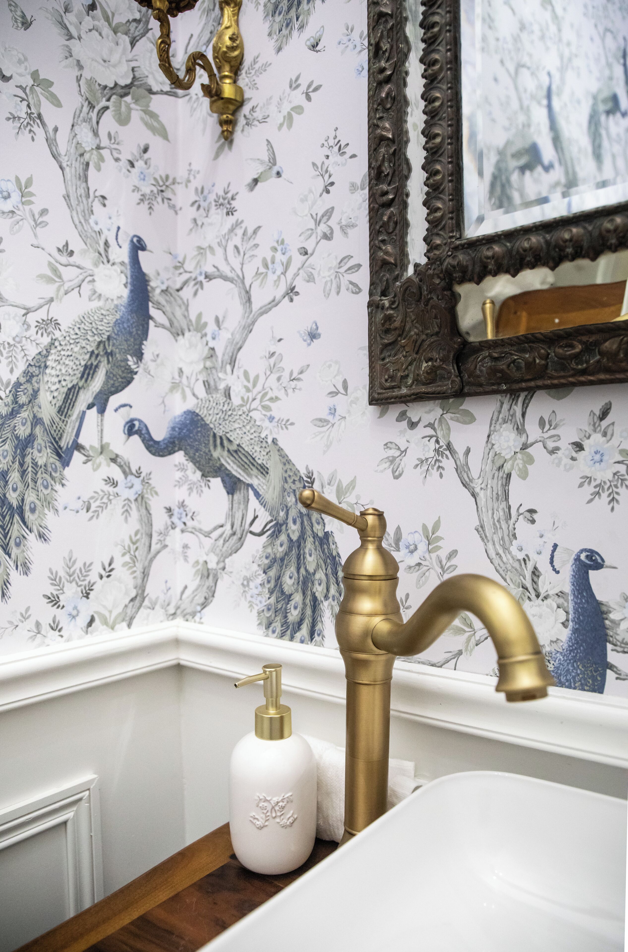 Un baño con un revestimiento decorativo de diseños de pavo real, un grifo dorado y un dispensador de jabón blanco.