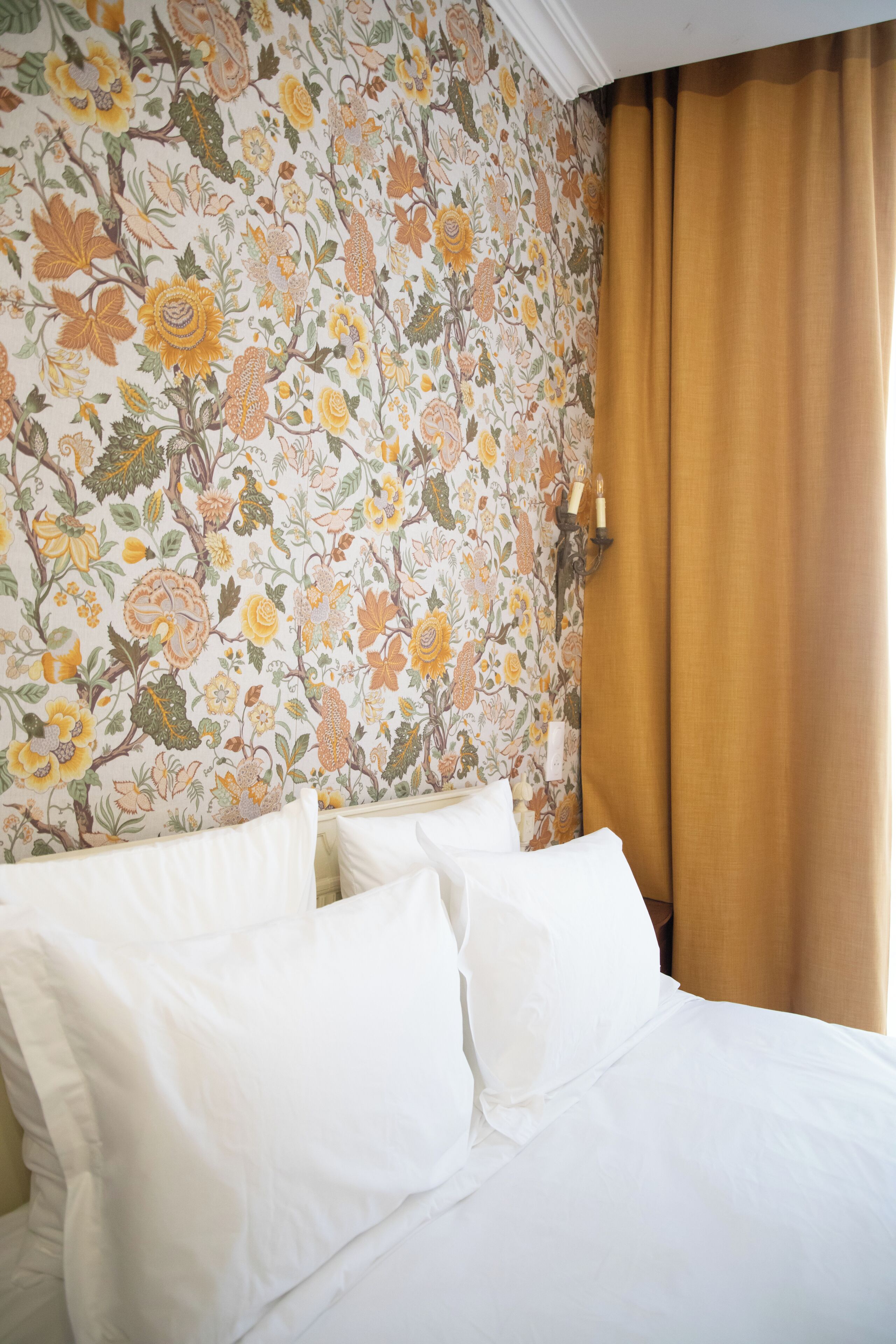 A hotel room with a floral wallpaper, a bed with white linens, and a window with a brown curtain.