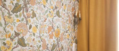 Une chambre d’hôtel avec un papier peint à motifs floraux, un lit avec du linge de maison blanc et une fenêtre ornée d’un rideau marron.