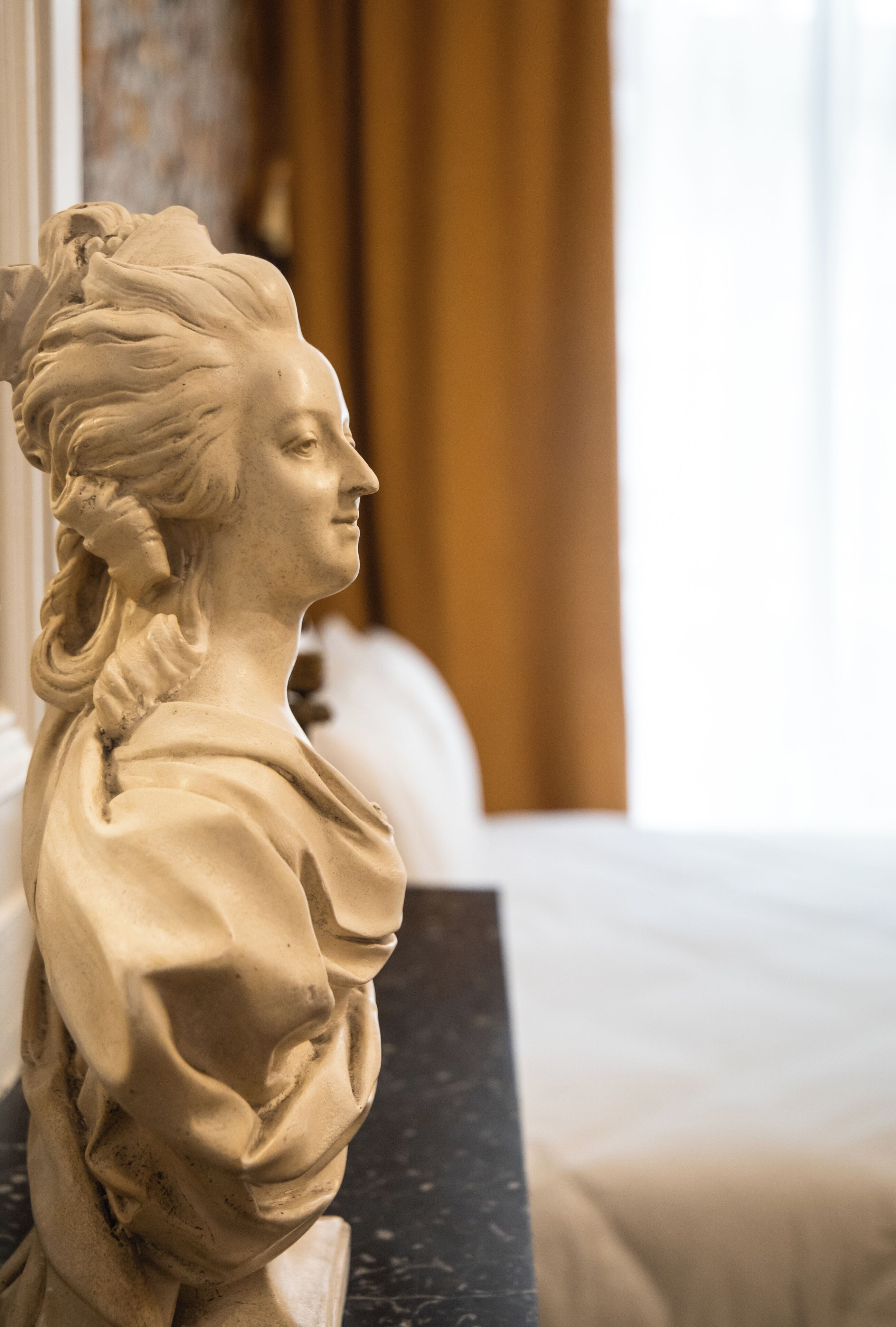 A bust sculpture on a black marble surface in a room with a bed and a window with curtains.