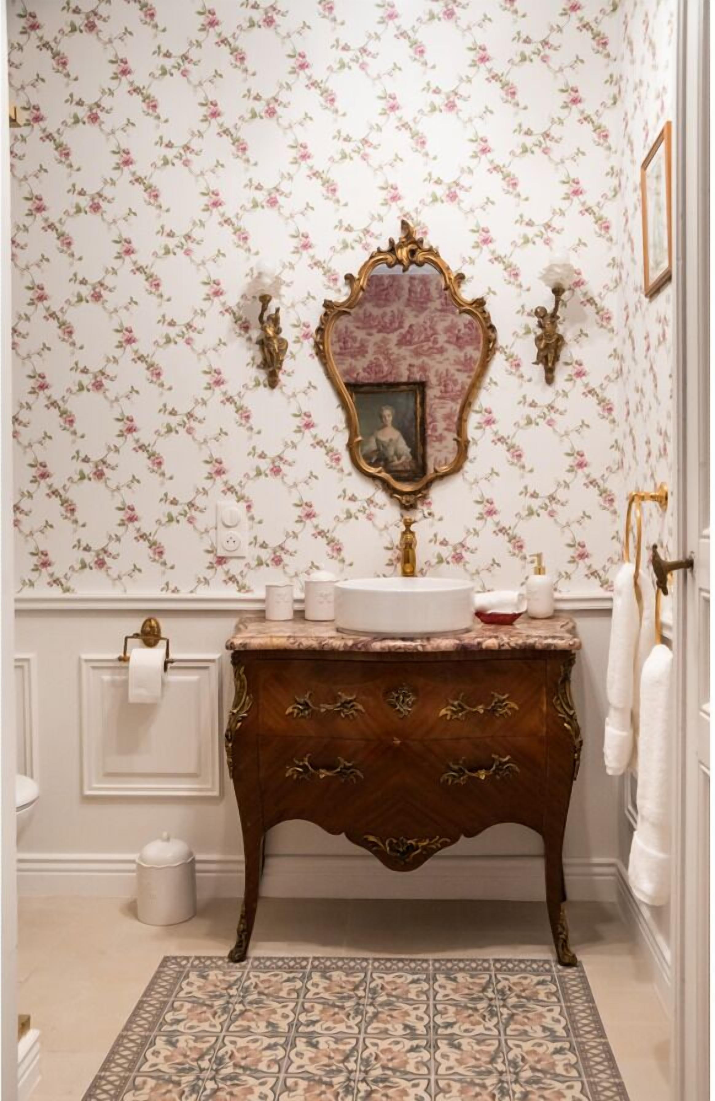 A bathroom with floral wallpaper, a marble sink, ornate gold-framed mirror, and a patterned rug.