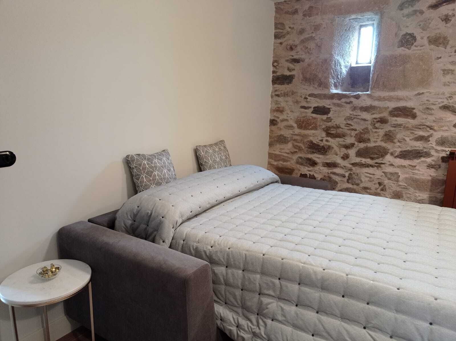 A bedroom with a stone wall, a bed with a quilted duvet, two patterned pillows, a small round table, and a chair.