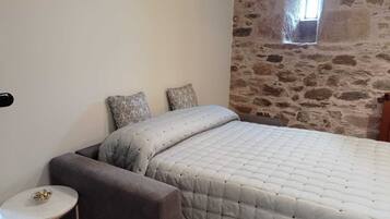 A bedroom with a stone wall, a bed with a quilted duvet, two patterned pillows, a small round table, and a chair.