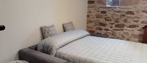 Un dormitorio con pared de piedra, una cama con edredón acolchado, dos almohadas estampadas, una mesita redonda y una silla.