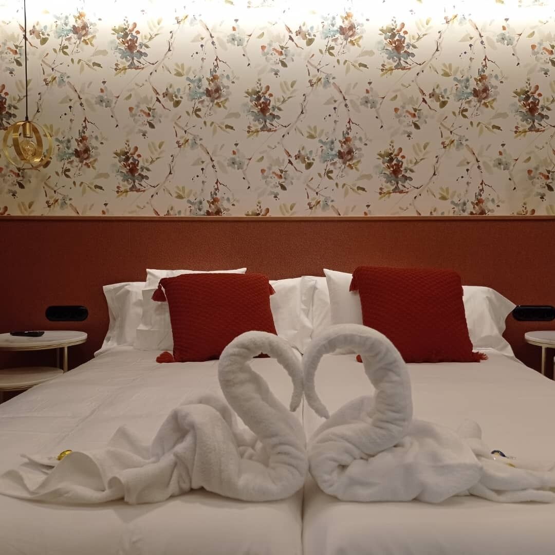 A hotel room with a floral wallpaper, a bed with white linens and red pillows, and a unique swan-shaped towel arrangement.