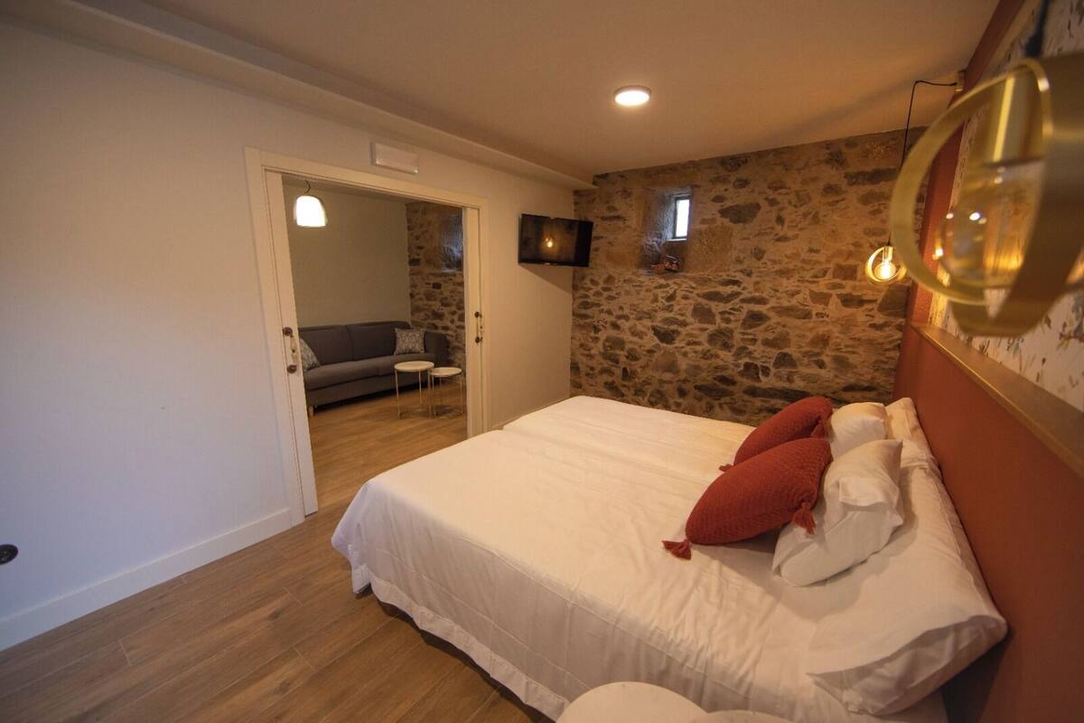 A bedroom with a stone wall, a bed with white bedding and red pillows, a small TV on the wall, and a wooden floor.