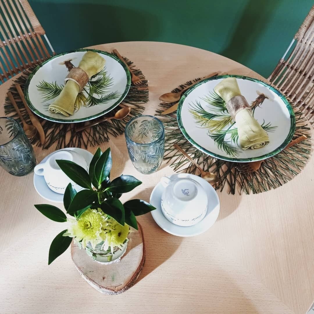 A round wooden table with two plates, each with a rolled-up napkin and a spoon, and a glass pitcher. There is a small potted plant with green leaves and yellow flowers on the table.