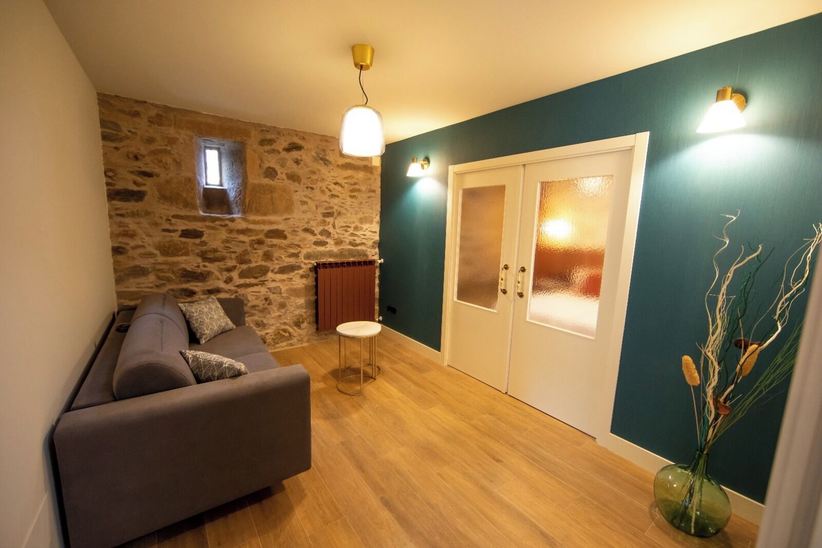 Un salon avec un mur en pierre, un canapé gris, une petite table ronde et une porte dont les panneaux sont en verre dépoli.