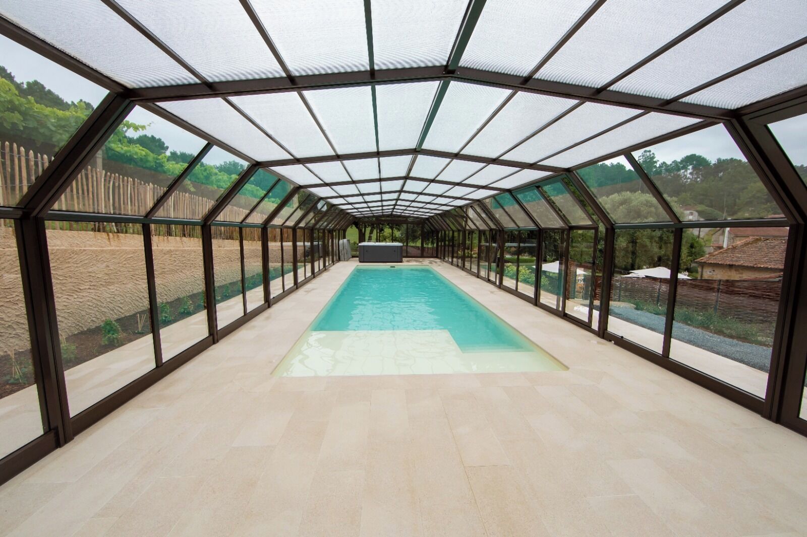 A covered swimming pool with a glass roof, surrounded by a fence, and a view of the countryside.