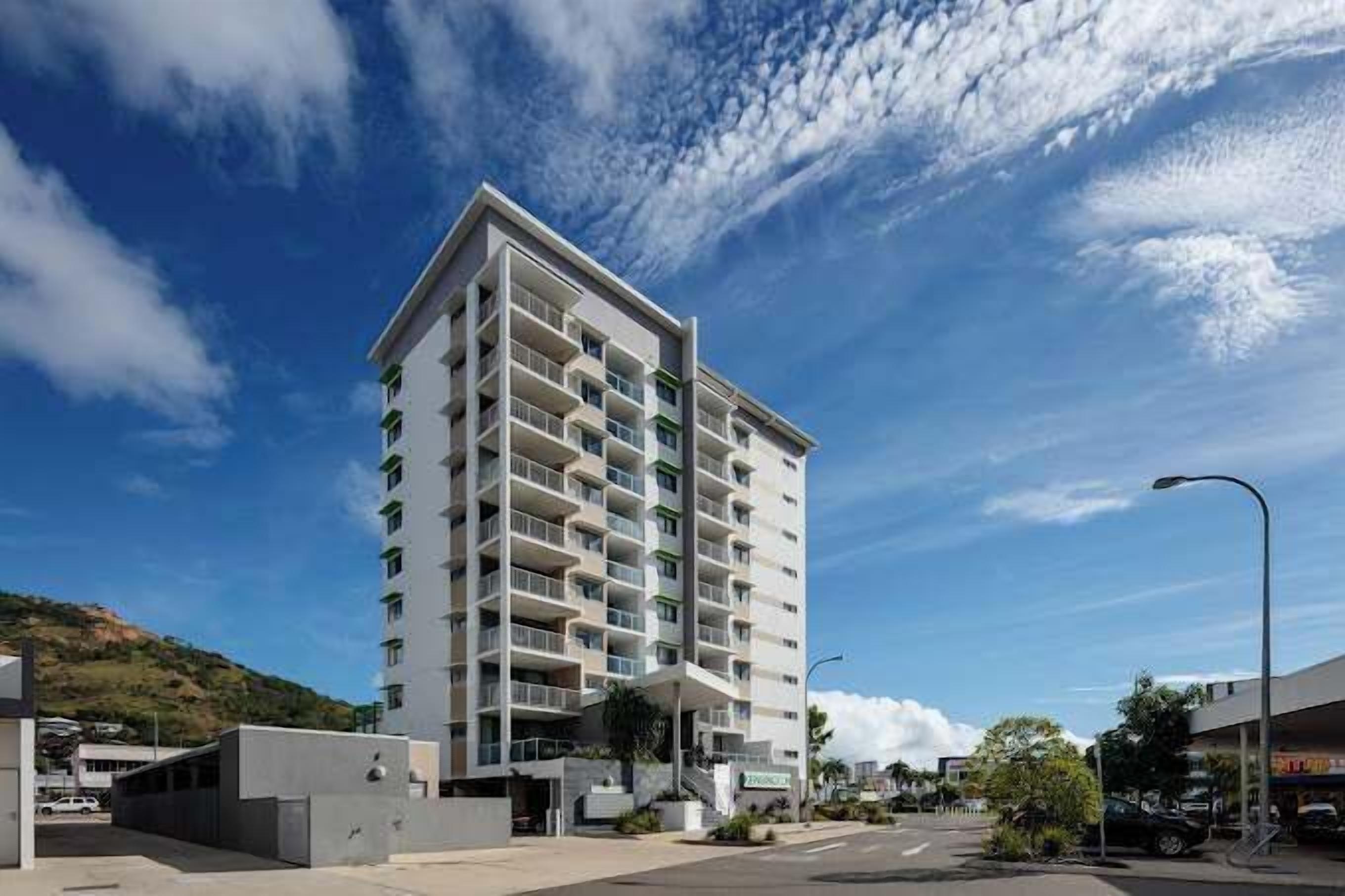 A modern multi-story hotel with balconies, a parking area, and a clear sky.