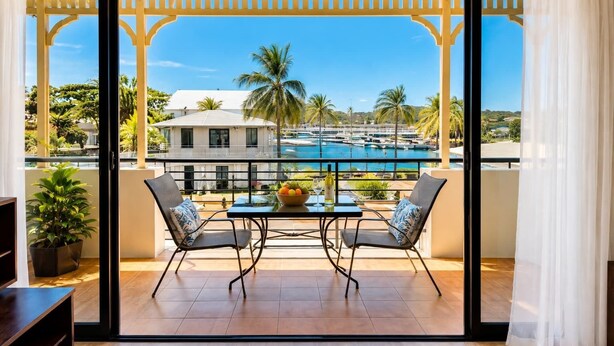 A balcony with a view of a pool, two chairs, a table with a bowl of fruit and a bottle, and a potted plant. - Anchorage Escape Townsville | Central Location & Marina Views (South Townsville)