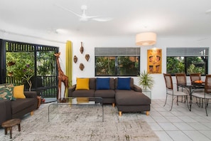 A modern living room with a sectional sofa, a dining table, and chairs. There is a large window with blinds, a ceiling fan, and a decorative giraffe sculpture. - North Ward Tropical Escape for 4 – Steps from the Beach & Close to CBD (North Ward)