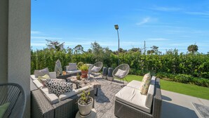 Un patio exterior con mobiliario de mimbre, una mesa con plantas en macetas y vistas a un jardín exuberante.