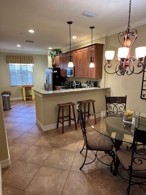 A kitchen with wooden cabinets, a stainless steel refrigerator, a microwave, and a sink. There is a dining area with a glass table and metal chairs. - Ole’ at Lely Resort. Adorable 2-bedroom casita, perfect for relaxing stays (Naples Fl.)