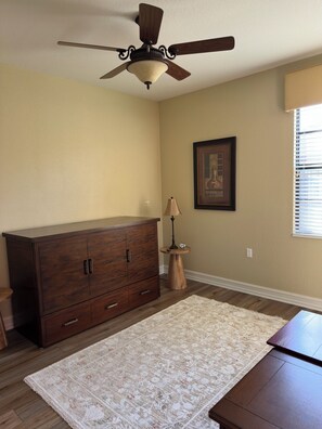 A room with a ceiling fan, a large wooden dresser, a small round table with a lamp, and a framed picture on the wall. - Ole’ at Lely Resort. Adorable 2-bedroom casita, perfect for relaxing stays (Naples Fl.)