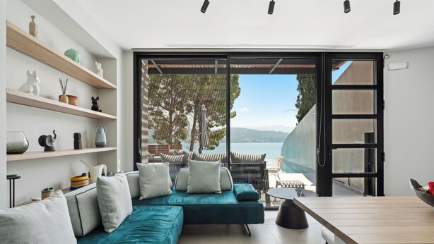 Ein modernes Wohnzimmer mit einem türkisfarbenen Sofa, weißen Kissen und einem Holztisch. Regale mit Dekorationselementen und Blick ins Freie.