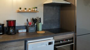 A compact kitchen with a dishwasher, oven, and a built-in stove. There are shelves with kitchen utensils and a coffee maker. - Le Rissas, gite for 4 _ Lacets Ventoux (Beaumont-du-Ventoux)